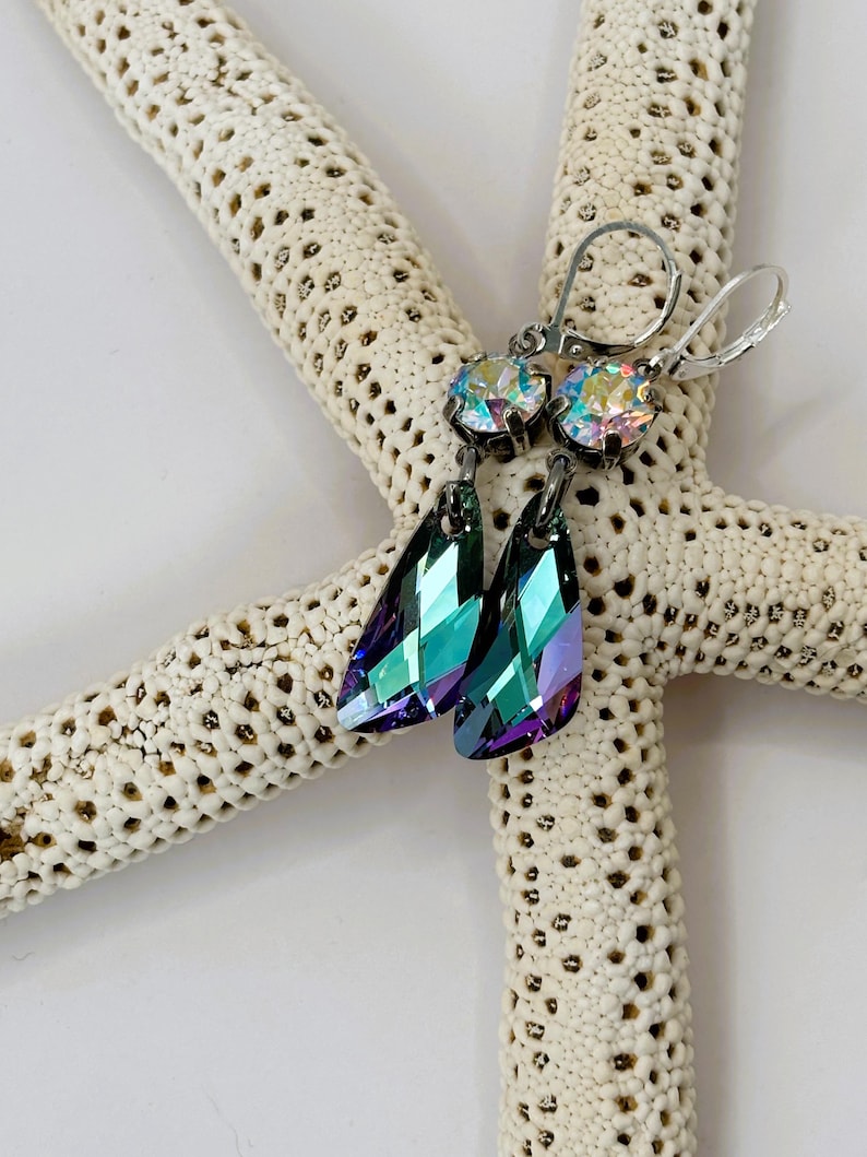 May include: A pair of silver-toned earrings with iridescent, teardrop-shaped crystals. The earrings have a lever-back closure and are displayed on a white starfish. The crystals reflect shades of teal, purple, and green.