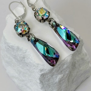 May include: A pair of silver-toned drop earrings. Each earring features a round, iridescent crystal at the top and a teardrop-shaped, multi-colored crystal pendant. The crystals reflect shades of blue, green, and purple.
