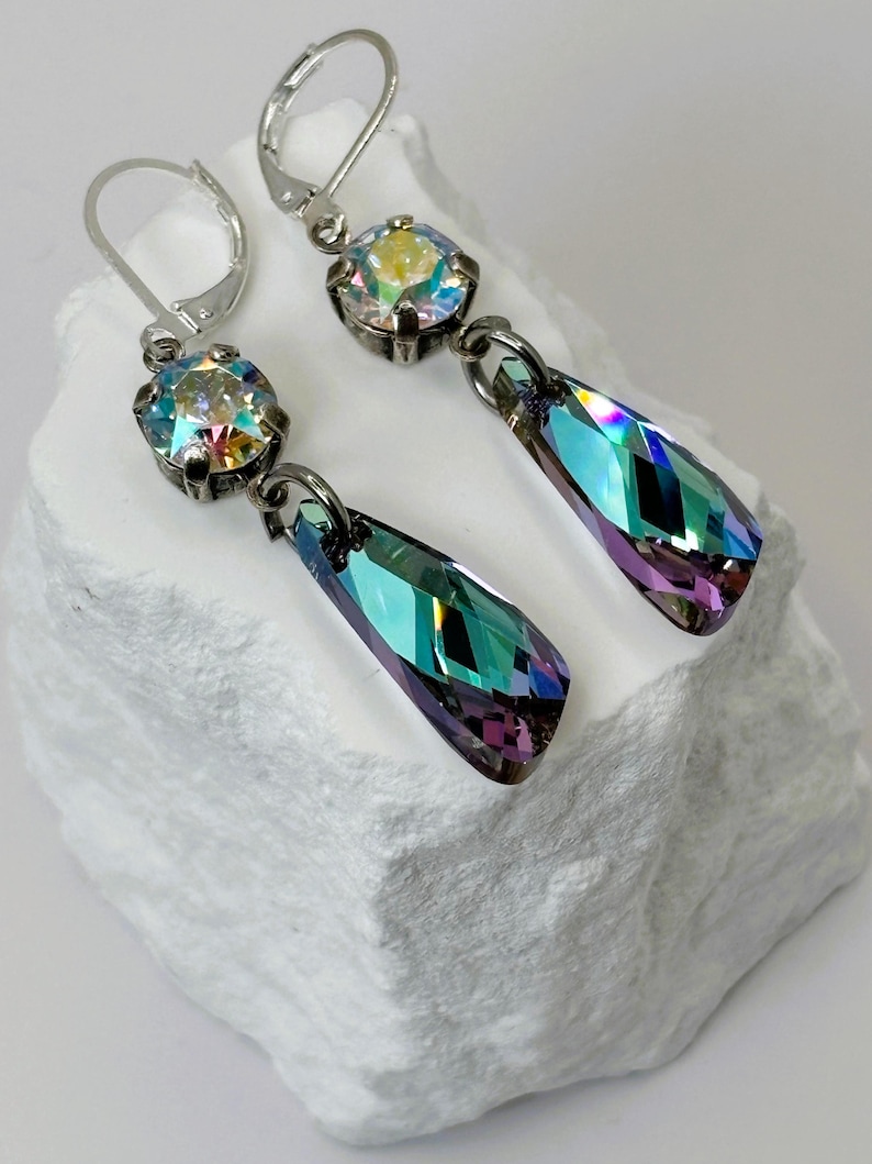 May include: A pair of silver-toned drop earrings. Each earring features a round, iridescent crystal at the top and a teardrop-shaped, multi-colored crystal pendant. The crystals reflect shades of blue, green, and purple.
