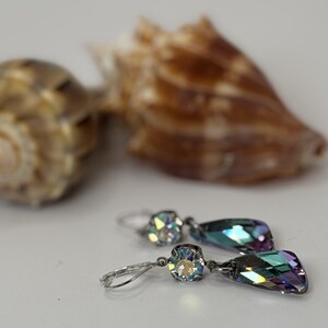 May include: A pair of silver drop earrings with iridescent, teardrop-shaped crystals. The earrings are displayed with two seashells in the background, creating a beachy aesthetic. The crystals reflect a range of colors.