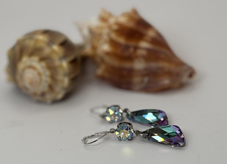 May include: A pair of silver drop earrings with iridescent, teardrop-shaped crystals. The earrings are displayed with two seashells in the background, creating a beachy aesthetic. The crystals reflect a range of colors.