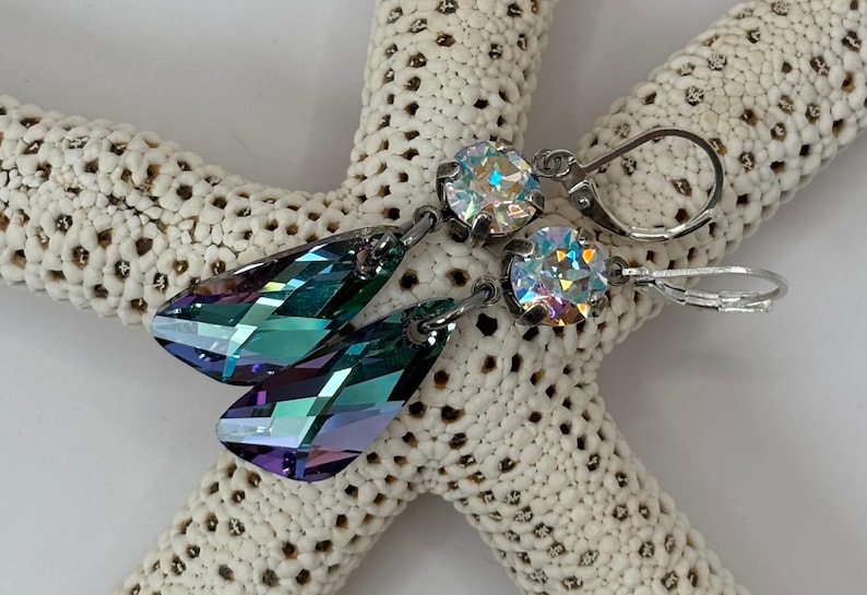 May include: A pair of earrings with iridescent, teardrop-shaped crystals and round, clear crystals. The earrings have silver-colored metal findings and are displayed on a white, textured starfish.