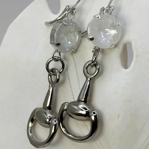 May include: A pair of silver-toned earrings. Each earring features a clear, round crystal above a horse bit charm. The earrings hang from a silver hook. The crystals have a faceted appearance, reflecting light. The horse bit charms have a detailed, realistic design.