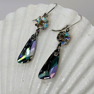 May include: A pair of dangle earrings with iridescent, teardrop-shaped crystals. Each earring features a smaller, round, iridescent crystal at the top, connected to a larger, faceted crystal. The earrings are set on silver-colored hooks, displayed on a white, textured surface.