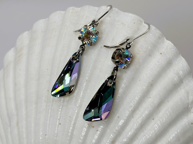 May include: A pair of dangle earrings with iridescent, teardrop-shaped crystals. Each earring features a smaller, round, iridescent crystal at the top, connected to a larger, faceted crystal. The earrings are set on silver-colored hooks, displayed on a white, textured surface.