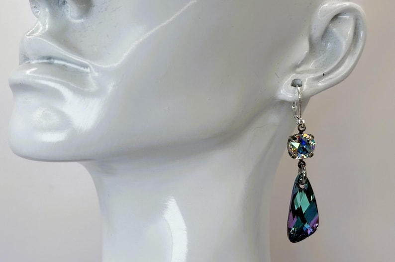 May include: A pair of dangle earrings featuring iridescent crystals. Each earring has a square-cut crystal at the top and a teardrop-shaped crystal below. The crystals display a range of colors, including green, blue, and purple, and are set in a silver-tone metal.