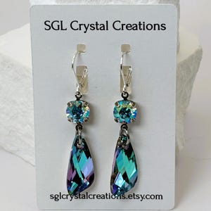 May include: A pair of dangle earrings with iridescent blue and purple teardrop-shaped crystals. Each earring has a round, iridescent blue crystal at the top, connected to a silver-tone lever back. The card reads "SGL Crystal Creations" and "sglcrystalcreations.etsy.com".