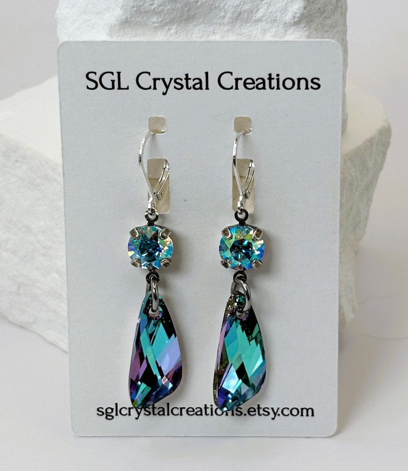 May include: A pair of dangle earrings with iridescent blue and purple teardrop-shaped crystals. Each earring has a round, iridescent blue crystal at the top, connected to a silver-tone lever back. The card reads "SGL Crystal Creations" and "sglcrystalcreations.etsy.com".