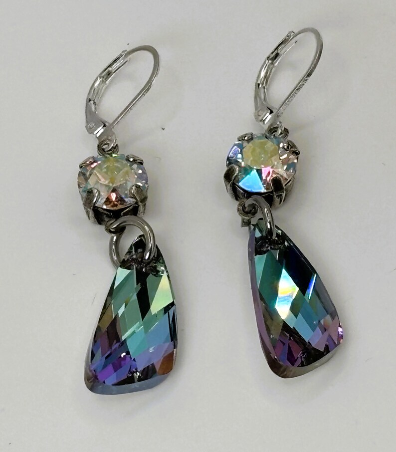 May include: A pair of silver-toned drop earrings. Each earring features a round, iridescent crystal at the top, connected to a teardrop-shaped, multi-colored crystal pendant. The pendants reflect shades of blue, green, and purple.
