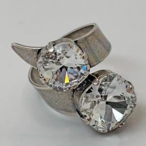 May include: A bypass ring crafted from a tarnished silver-toned metal, featuring two large, round, clear faceted stones set in prong settings. The band has a wide, flat profile that wraps around the finger, showcasing a metallic finish.