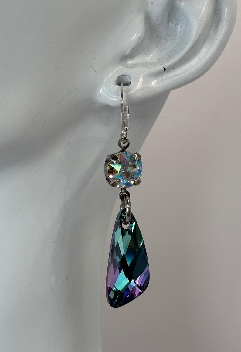May include: A single, elegant earring with a silver-tone hook. The earring features a round, iridescent crystal above a larger, teardrop-shaped crystal with shades of teal, purple, and green. Jewelry.