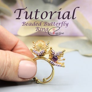 Könnte beinhalten: Ein goldener Ring mit zwei Perlen-Schmetterlingsanhängern. Ein Schmetterlingsanhänger ist mit einem hellgelben Edelstein verziert, der andere mit einem violetten Edelstein.