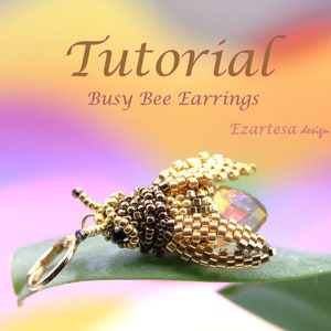 Può includere: Orecchini a forma di ape in perline dorate e marroni con un accento di cristallo trasparente. Gli orecchini sono appesi a una foglia verde. Il testo "Tutorial Busy Bee Earrings Ezarteasa design" è visibile nella parte superiore dell'immagine.