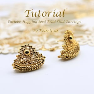 Puede incluir: Un par de pendientes de botón de cuentas de semillas doradas que abrazan el lóbulo de la oreja. Los pendientes están hechos de pequeñas cuentas doradas y tienen forma de corazón.