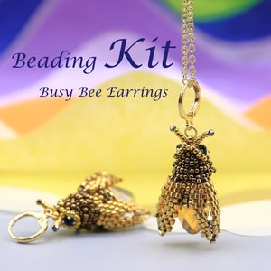 May include: A close-up of a pair of beaded bee earrings. The earrings are gold and brown, with black accents for the eyes. The text "Beading Kit - Busy Bee Earrings" is in the background. One earring is hanging from a gold chain.
