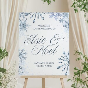 May include: A wedding welcome sign on a wooden easel. The sign is white with blue snowflake accents and text that reads "Welcome to the Wedding of Elsie & Noel" with the date January 10, 2026. The sign is surrounded by greenery and white flowers.