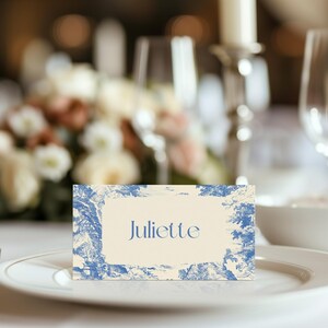 Puede incluir: Una tarjeta de sitio con el nombre "Juliette" en escritura azul sobre fondo crema, enmarcada por un dise&ntilde;o toile de Jouy azul y blanco. La tarjeta est&aacute; colocada sobre un plato blanco, con un tenedor a la izquierda, para una mesa elegante.