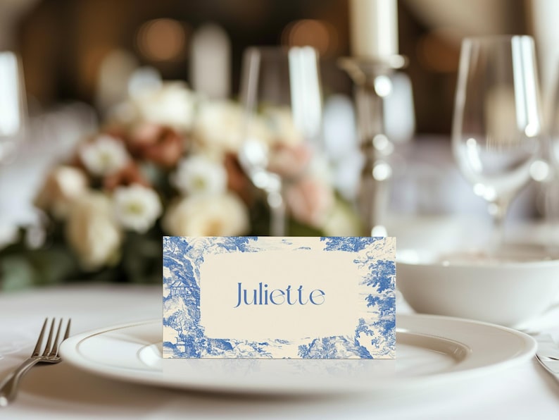 Puede incluir: Una tarjeta de sitio con el nombre "Juliette" en escritura azul sobre fondo crema, enmarcada por un dise&ntilde;o toile de Jouy azul y blanco. La tarjeta est&aacute; colocada sobre un plato blanco, con un tenedor a la izquierda, para una mesa elegante.