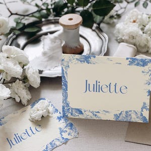 Puede incluir: Dos tarjetas de mesa con el nombre "Juliette" en escritura azul, enmarcadas por un dise&ntilde;o de toile de Jouy azul y blanco. Las tarjetas est&aacute;n sobre una mesa con flores blancas, una bandeja plateada y una bobina de madera.