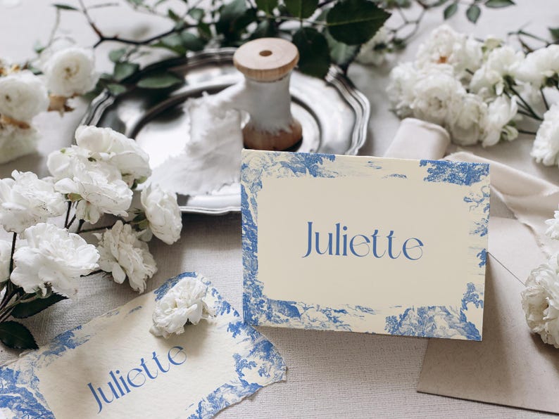 Puede incluir: Dos tarjetas de mesa con el nombre "Juliette" en escritura azul, enmarcadas por un dise&ntilde;o de toile de Jouy azul y blanco. Las tarjetas est&aacute;n sobre una mesa con flores blancas, una bandeja plateada y una bobina de madera.