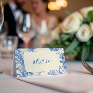 Puede incluir: Una tarjeta de mesa plegada con el nombre "Juliette" en elegante escritura azul. La tarjeta presenta un borde de dise&ntilde;o toile de Jouy azul y blanco. La escena est&aacute; ambientada sobre un mantel blanco, con un fondo borroso de una recepci&oacute;n de boda.
