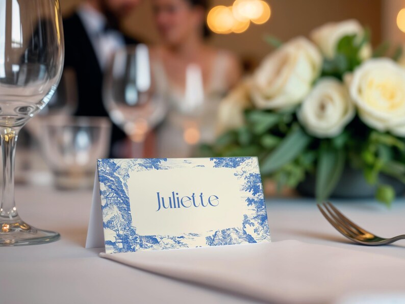 Puede incluir: Una tarjeta de mesa plegada con el nombre "Juliette" en elegante escritura azul. La tarjeta presenta un borde de dise&ntilde;o toile de Jouy azul y blanco. La escena est&aacute; ambientada sobre un mantel blanco, con un fondo borroso de una recepci&oacute;n de boda.