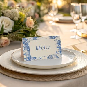 Puede incluir: Un lugar en la mesa con una tarjeta con el nombre "Juliette" en azul, sobre un plato blanco con borde dorado. La tarjeta tiene un dise&ntilde;o azul y blanco. Un arreglo floral y copas de vino son visibles.