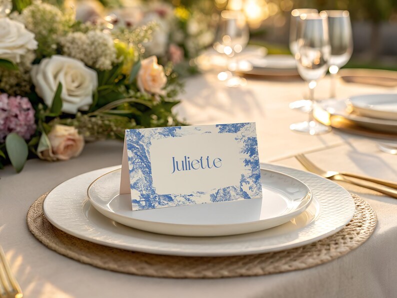 Puede incluir: Un lugar en la mesa con una tarjeta con el nombre "Juliette" en azul, sobre un plato blanco con borde dorado. La tarjeta tiene un dise&ntilde;o azul y blanco. Un arreglo floral y copas de vino son visibles.