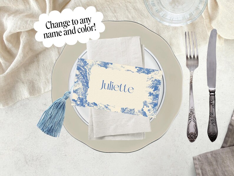 Puede incluir: Un montaje de mesa con una tarjeta con el nombre "Juliette". La tarjeta tiene un dise&ntilde;o toile de Jouy azul y blanco y una borla azul. Un tenedor y un cuchillo est&aacute;n a la derecha del plato. El texto "&iexcl;Cambia a cualquier nombre y color!" est&aacute; en una burbuja.