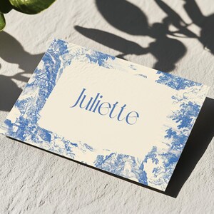 Puede incluir: Una tarjeta rectangular con el nombre "Juliette" en escritura azul. La tarjeta tiene un centro color crema y un borde toile azul y blanco. La tarjeta est&aacute; sobre una superficie texturizada, gris claro, con sombras de follaje.