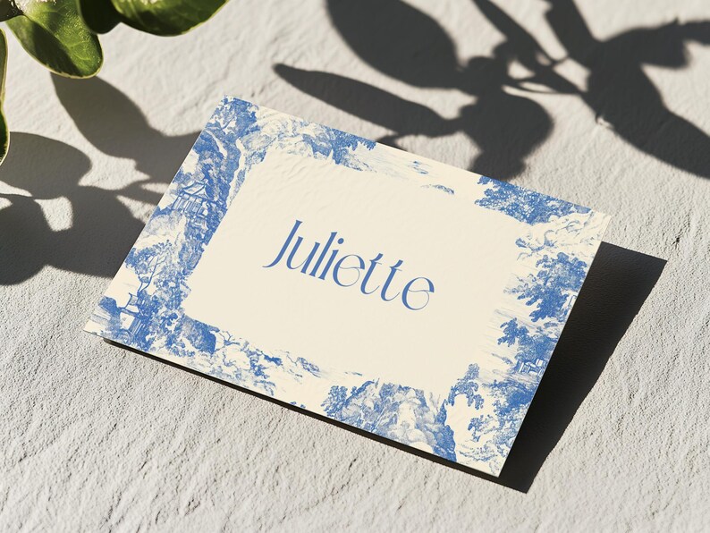Puede incluir: Una tarjeta rectangular con el nombre "Juliette" en escritura azul. La tarjeta tiene un centro color crema y un borde toile azul y blanco. La tarjeta est&aacute; sobre una superficie texturizada, gris claro, con sombras de follaje.