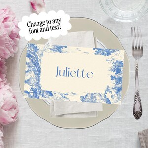 Puede incluir: Un cubierto con una tarjeta de sitio de estilo toile de Jouy azul y blanco que dice "Juliette". La tarjeta est&aacute; sobre una servilleta blanca, un plato gris claro y junto a un tenedor y un cuchillo. El texto "&iexcl;Cambia a cualquier fuente y texto!" est&aacute; en una burbuja.