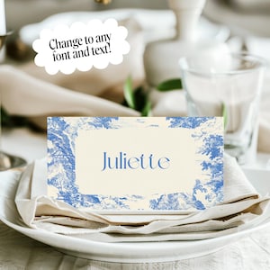 Puede incluir: Una tarjeta de sitio con el nombre "Juliette" en escritura azul, colocada sobre un plato blanco con una servilleta. La tarjeta tiene un fondo crema con un borde azul toile de Jouy. Tambi&eacute;n se ven una vela y cubiertos.