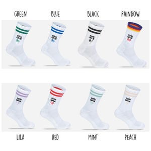 Op de afbeelding: Acht paar witte sokken met gekleurde strepen en tekst. De sokken hebben groene, blauwe, zwarte, regenboog-, lila, rode, mintgroene en perzikstrepen. Elke sok heeft de tekst "BOOK GIRLIE" en een strik-afbeelding.