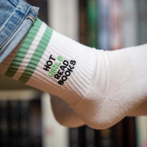 Könnte beinhalten: Weiße gerippte Socken mit grünen Streifen und dem gestickten Text "HOT GIRLS READ BOOKS" in Grün und Schwarz. Die Socken werden mit einer blauen Jeans getragen.