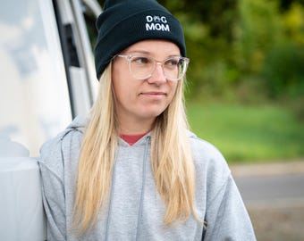 Op de afbeelding: Een donkergroene beanie met de tekst "DOG MOM" in het wit, gecombineerd met een bril met helder montuur. De persoon draagt een grijze hoodie met een rood ondershirt.
