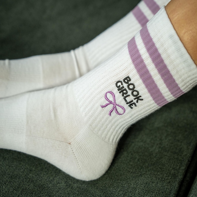 Op de afbeelding: Witte sokken met lavendelkleurige strepen en geborduurde tekst en strik. De tekst luidt "BOOK GIRLIE" in zwart, met een lavendelkleurige strik eronder. De sokken zijn geribbeld en lijken van een zacht materiaal te zijn.