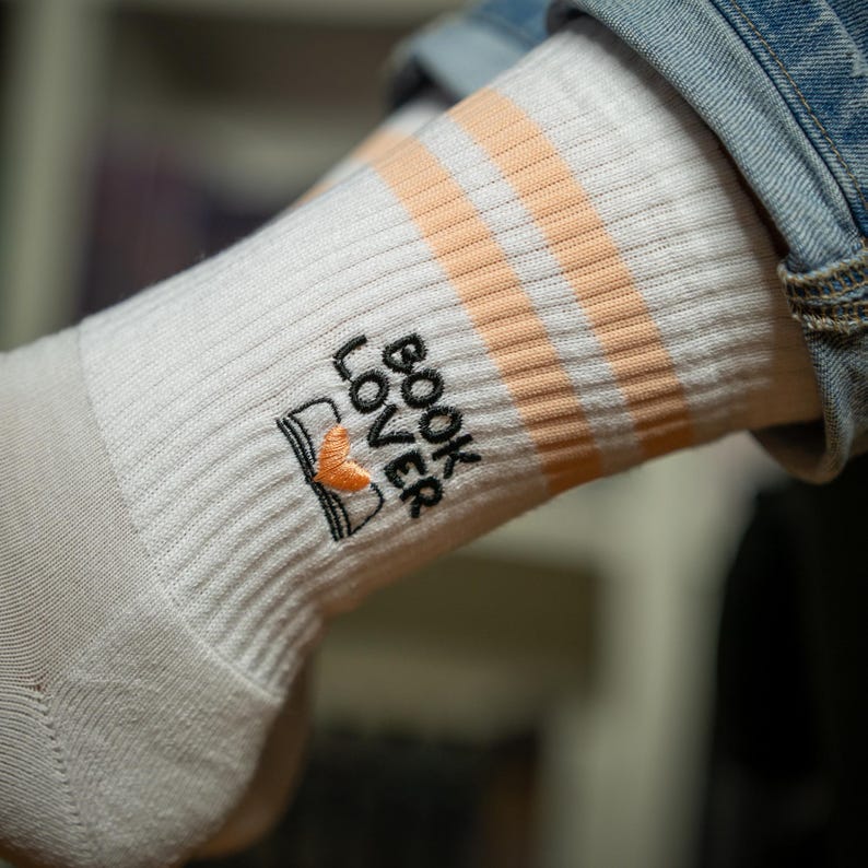 May include: White ribbed socks with peach-colored stripes and the embroidered text "BOOK LOVER" with a book graphic. The socks are visible on a person's leg, with a denim jacket sleeve in the background.
