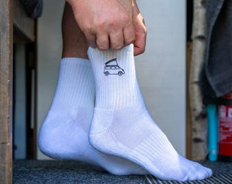 Op de afbeelding: Witte sportsokken met een geribbelde textuur. Elke sok heeft een klein, geborduurd zwart camperbusje-ontwerp bij de manchet. De sokken worden op de voeten van een persoon omhoog getrokken.