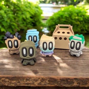 May include: A selection of brightly coloured, stylised figurines with large oval eyes and geometric features. The figures are in various colours, including tan, blue, green, and beige, and are arranged on a wooden surface. A small cardboard box with a handle is also present.