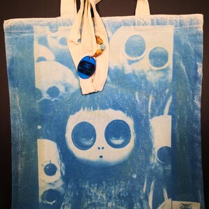 May include: A blue tote bag with a white strap and a blue and white image of a girl with large eyes. The bag has a small blue tag with the text "NOCTURNAL INN" on it.