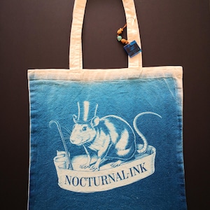 May include: A blue tote bag featuring a white rat wearing a top hat and holding a cane. The rat is perched on a white banner with the text "Nocturnal-Ink". The bag has a blue and white ombre effect and a small logo on the bottom right corner that reads "Nocturnal Ink".