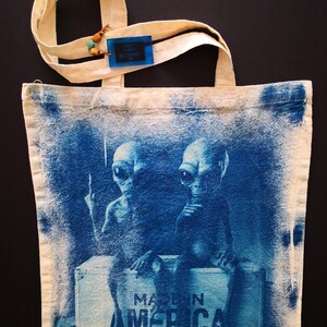 May include: A white canvas tote bag with blue alien figures printed on it. The aliens are sitting on a box that says "Made in America".