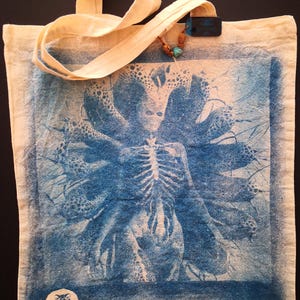 May include: A white canvas tote bag featuring a blue and white image of a skeletal figure with multiple arms. The image is surrounded by a white border. The bag has a blue and white logo that says "Nocturnal Ink" in the bottom left corner.