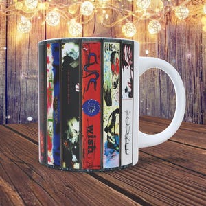 May include: White ceramic mug featuring album art from The Cure. The mug displays a collage of album covers in various colours, including red, blue, and black. The mug has a white handle and is set on a wooden surface.