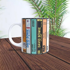 May include: White ceramic mug featuring a design of stacked cassette tapes with David Bowie album covers. The album titles include "Stage", "Never Let Me Down", "Ziggy Stardust", "Let's Dance", and "Pin Ups".