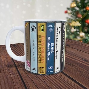 May include: White ceramic mug featuring a design of vintage cassette tapes with Elvis Presley album covers. The cassette tapes include "Elvis Presley - Blue Suede Shoes", "Elvis in G.I. Blues", "Elvis' Christmas Album", "Elvis - Are You Lonesome Tonight", and "Elvis - TV Special".