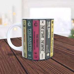 May include: White ceramic mug featuring a design of stacked cassette tapes with The Smiths band albums. The cassette tapes are labelled with album titles such as "Meat Is Murder", "Strangeways, Here We Come", "The 12" Tape", "The World Won't Listen", "I Started Something I Couldn't Finish", and "Ask".