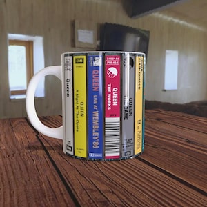 May include: A white coffee mug featuring a design of six cassette tapes with Queen album covers. The tapes are arranged vertically around the mug. The album covers include "Queen Live at Wembley '86", "The Works", "Queen II", "A Night at the Opera", "Flash Gordon", and "Queen".