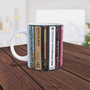May include: White ceramic mug featuring a design of stacked cassette tapes with Depeche Mode album covers. The album titles include "Songs of Faith and Devotion", "Personal Jesus", "Black Celebration", "Music for the Masses", and "Never Let Me Down Again".
